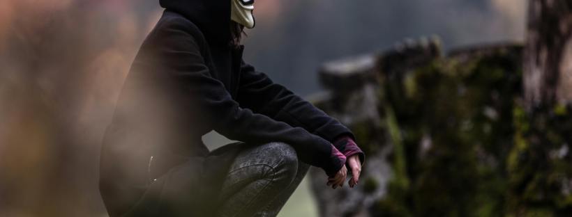 A person wearing a dark hooded coat and a Guy Fawkes mask squats on a mossy tree stump in a forested area. The scene is misty and somber, with blurred foreground elements and muted autumn colors in the background. The figure’s posture is contemplative and still, evoking a sense of solitude or quiet resistance.