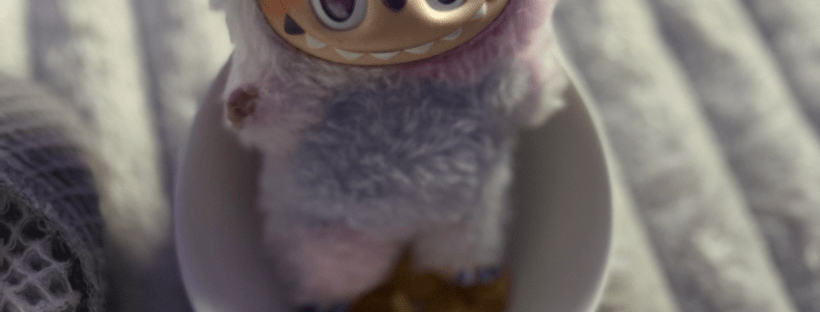 A Labubu figure dressed in a fluffy pastel blue and purple costume with bunny ears sits inside a small white bowl filled with snack pieces, resting on a soft gray blanket.
