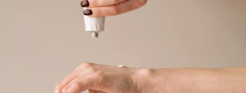 Close-up of two pale hands with dark polished nails. One hand squeezes lotion from a white tube onto the other hand’s wrist. (Yes… it puts the lotion on the skin.)