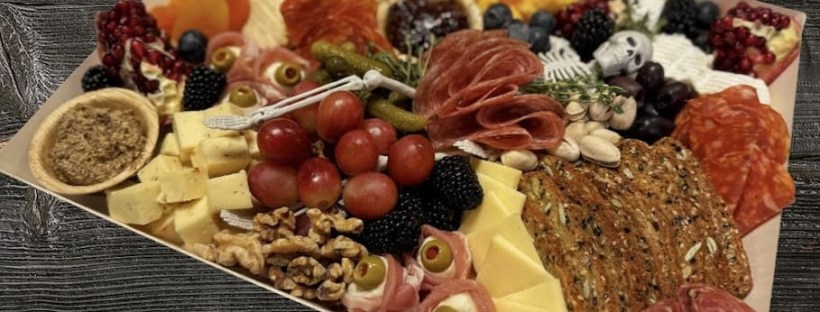 A Halloween-themed charcuterie board arranged with layers of meats, cheeses, crackers, fruit, nuts, and spooky touches like tiny skull decorations and olive “eyeballs.” The display includes blackberries, grapes, pomegranate seeds, and bright orange cheese cubes, all set against a dark wood background.
