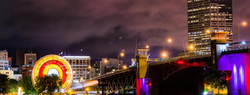 Nighttime view of downtown Portland, Oregon, featuring the Hawthorne Bridge lit with purple and red lights and a glowing Ferris wheel spinning near the waterfront. The city skyline rises behind the bridge under a cloudy, pink-tinted sky, with reflections of the lights shimmering on the Willamette River.
