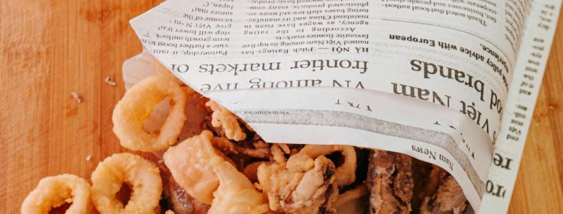 Fried calamari spilling out of a cone made from newspaper onto a wooden cutting board, with two whole lemons resting nearby.