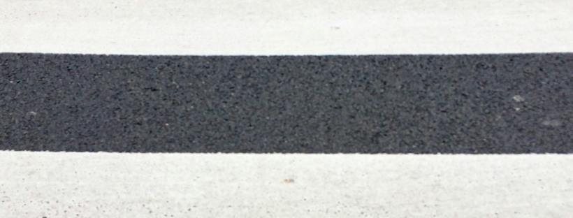 Close-up view of a pedestrian crosswalk, showing alternating wide white painted stripes and dark asphalt bands stretching horizontally across the frame.
