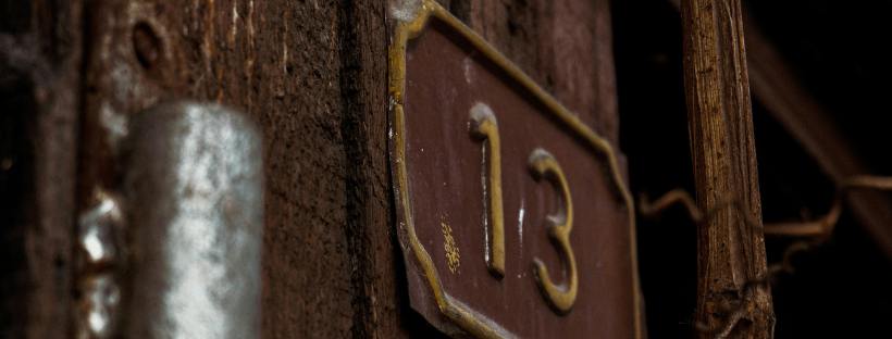 Close-up of an aged wooden structure with a metal number “13” affixed to it, surrounded by branches and deep shadow, highlighting rough textures and a quiet, moody atmosphere.