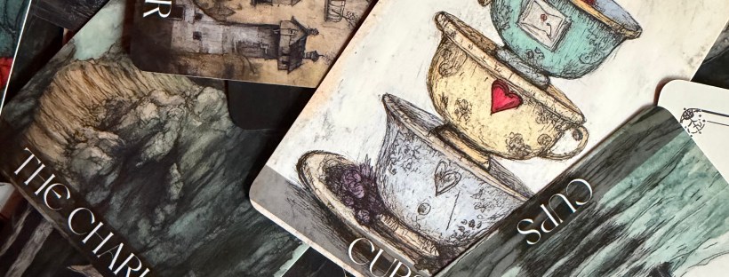 A scattered pile of tarot cards from the Daydream Tarot deck, showing richly illustrated cards including The Tower, The Chariot, and multiple cards from the suit of Cups. The artwork is detailed and moody, rendered in muted tones with pops of color. The Five of Cups card is most prominent, depicting a whimsical stack of mismatched teacups in yellow, red, and teal.
