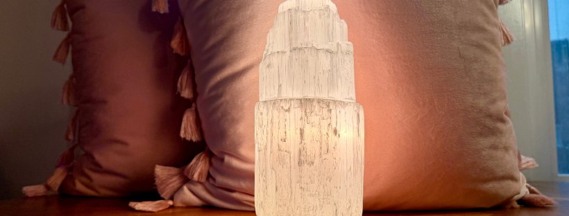 A glowing selenite tower lamp sits on a warm wood table, backlit by a window. Three tarot cards are laid face-down in front of it. Two dusty pink velvet pillows with tassel trim lean against the window behind it.
