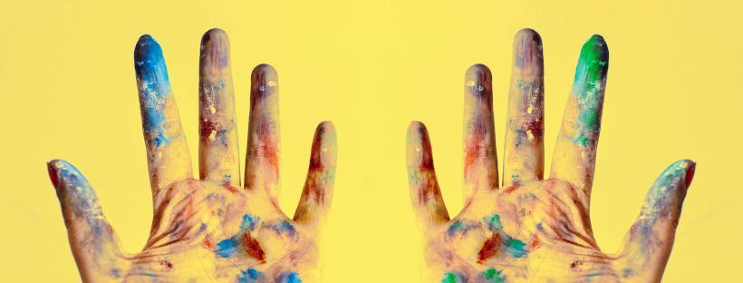 Two paint-covered hands held open, palms facing outward against a bright yellow background. The hands are smeared with blue, green, red, and brown paint across the fingers and palms.