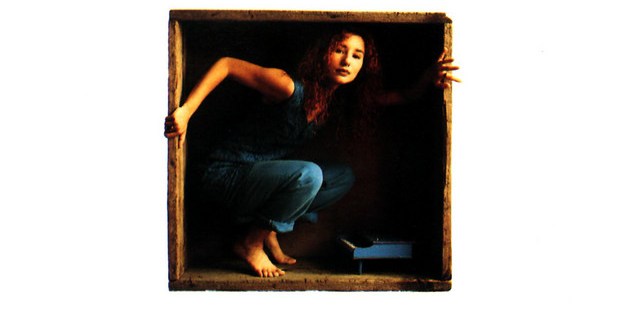 Album cover for Little Earthquakes by Tori Amos. White background with "TORI AMOS" in large weathered wood-block letters across the top. Centered in the middle of the cover, a small square box contains a photo of a young woman with long curly red hair, crouching barefoot in a confined wooden space, wearing a blue outfit, with a tiny toy piano at her feet. She looks directly at the camera. The album title Little Earthquakes appears in small italic text below the box.