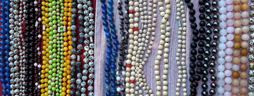 ⏺ Dozens of hand-tied beaded necklaces laid out side by side — semi-precious stone malas in every color. Greens, blues, amber, white, black, coral, turquoise, yellow.