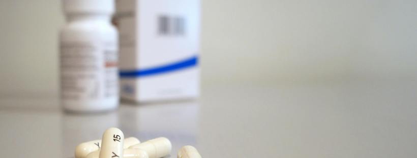 White medication capsules scattered on a pale surface, with a pill bottle and its packaging blurred in the background.