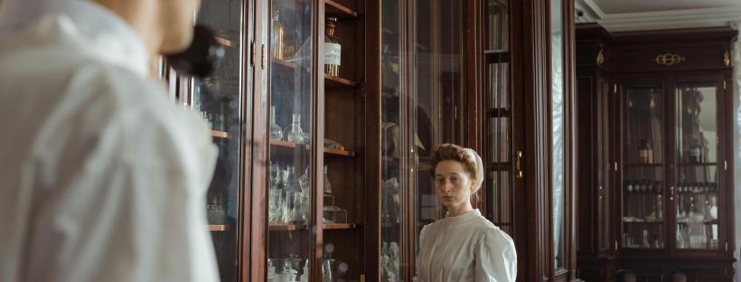 Vintage interior with woman in white dress examining wooden cabinets, evoking a historical apothecary setting.