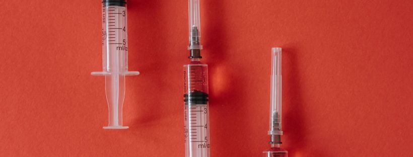 Three plastic syringes filled with red liquid, arranged diagonally across a deep coral background.