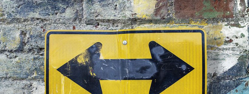 A scuffed yellow road sign with two black arrows pointing in opposite directions — one left, one right — mounted on a weathered brick wall with peeling paint in yellow, blue, red, and black.