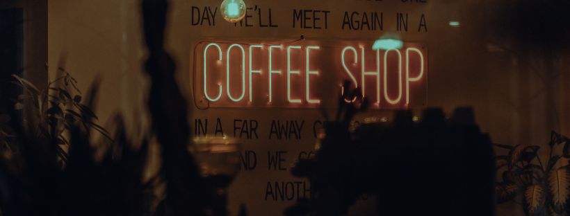 A dimly lit coffee shop interior at night. A cluster of bare Edison bulbs on black cords hangs from the ceiling, glowing pale blue-green. Behind them, a wall bears hand-lettered black text — "WE MET AT THE WRONG TIME. THAT'S WHAT I KEEP TELLING MYSELF ANYWAY. MAYBE ONE DAY WE'LL MEET AGAIN IN A —" — with a pink-and-white neon sign reading "COFFEE SHOP" mounted mid-wall. Silhouetted plants and an espresso machine sit in shadow in the foreground.